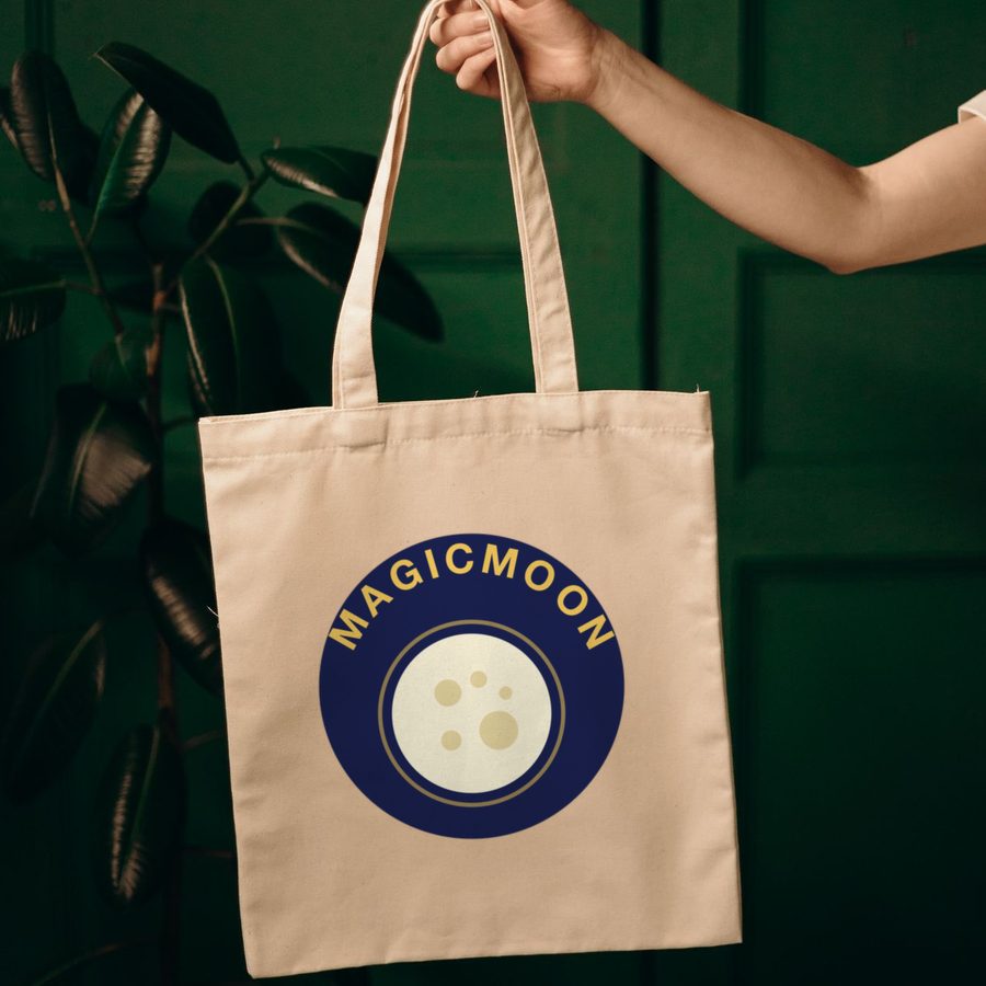 Hand holding a canvas tote bag with a full moon design and the word MAGICMOON curved around it printed on the front